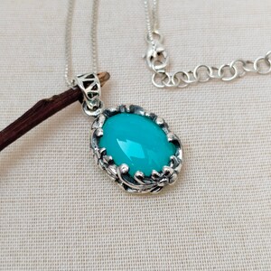 May include: A silver chain necklace with a turquoise oval pendant. The pendant is set in a silver frame with intricate floral designs.