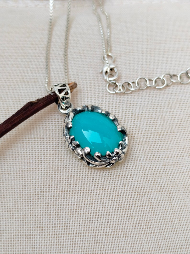May include: A silver chain necklace with a turquoise oval pendant. The pendant is set in a silver frame with intricate floral designs.