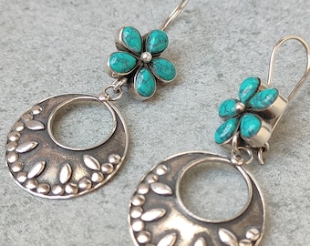May include: A pair of silver earrings featuring turquoise flower studs and circular pendants. The pendants are adorned with a pattern of small beads and leaf-shaped accents. The earrings have a hook closure.
