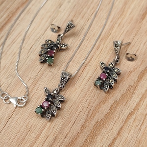 May include: A silver necklace and earrings set with a floral design featuring green and red gemstones. The pendant and earrings are adorned with marcasite.