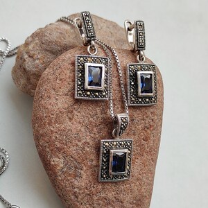Peut inclure: Un ensemble de collier et boucles d'oreilles en argent avec une pierre pr&eacute;cieuse bleue et des accents de marcassite. Le pendentif et les boucles d'oreilles sont de forme carr&eacute;e avec un design d&eacute;taill&eacute;.