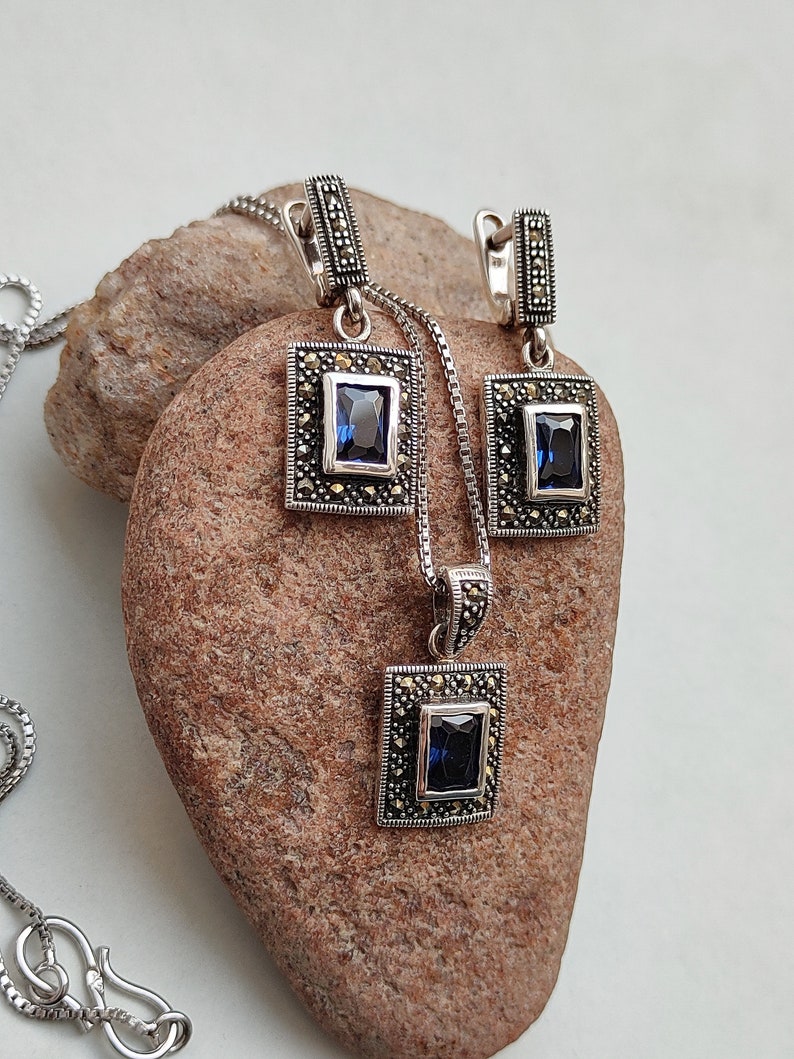 Peut inclure: Un ensemble de collier et boucles d'oreilles en argent avec une pierre pr&eacute;cieuse bleue et des accents de marcassite. Le pendentif et les boucles d'oreilles sont de forme carr&eacute;e avec un design d&eacute;taill&eacute;.