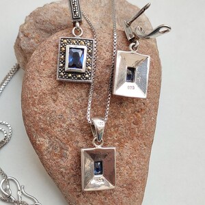 Peut inclure: Un collier en argent avec un pendentif carr&eacute; orn&eacute; d'une pierre pr&eacute;cieuse bleue entour&eacute;e de pierres de marcassite. Le collier est assorti &agrave; des boucles d'oreilles de m&ecirc;me design.