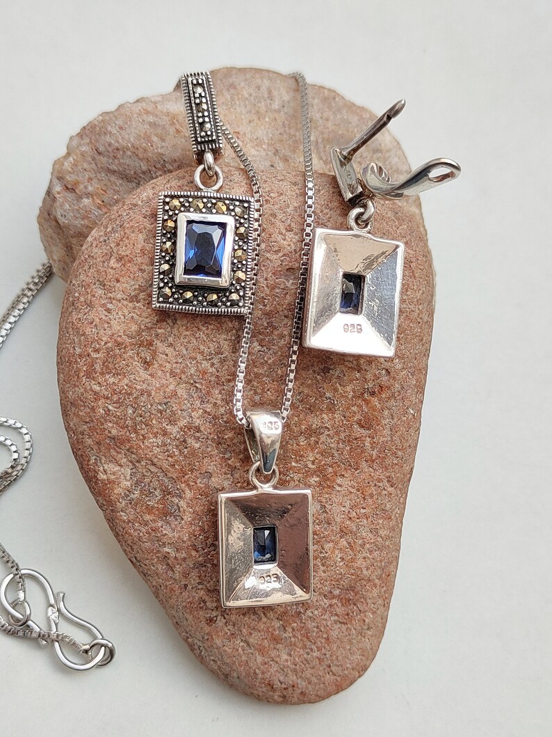 Peut inclure: Un collier en argent avec un pendentif carr&eacute; orn&eacute; d'une pierre pr&eacute;cieuse bleue entour&eacute;e de pierres de marcassite. Le collier est assorti &agrave; des boucles d'oreilles de m&ecirc;me design.