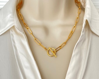 May include: A gold-colored chain necklace with an oval link design and a toggle clasp. The necklace is displayed on a mannequin wearing a white satin collared shirt. The necklace is a stylish accessory.