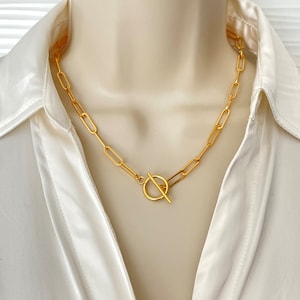 Op de afbeelding: Een goudkleurige ketting met een ovaal schakelontwerp en een tuimelsluiting. De ketting wordt getoond op een mannequin die een wit satijnen overhemd draagt. De ketting is een stijlvol accessoire.