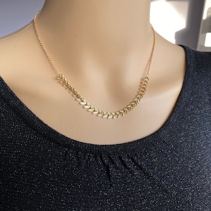 Könnte beinhalten: Eine goldene Halskette mit einem zarten Blattdesign. Die Kette wird von einer Person mit einem schwarzen Oberteil getragen.