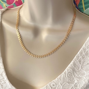 Könnte beinhalten: Eine goldene Halskette mit einem zarten Blattketten-Design. Die Halskette wird von einem Model mit einem weißen Spitzenoberteil getragen.