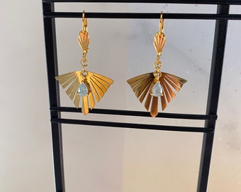 May include: A pair of gold-colored earrings with a fan-like design. Each earring features a small, light blue gemstone suspended below the fan. The earrings are displayed on a black metal stand.