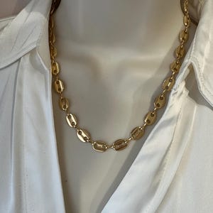 Op de afbeelding: Een goudkleurige ketting met een reeks ovaalvormige schakels. De ketting wordt getoond op een mannequin die een wit overhemd met kraag draagt. De schakels zijn gelijkmatig verdeeld en hebben een gepolijste afwerking, die licht reflecteert.