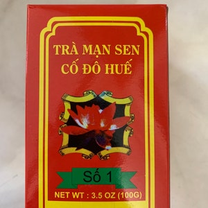 May include: Red box with yellow trim and a green label. The box contains a tea product called "Trà Man Sen Cố Đô Huế" with the number "Số 1" on the label. The text "NET WT: 3.5 OZ (100G)" and "HƯƠNG VỊ HẢO HẠNG" are also on the label.