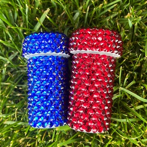 May include: Two bedazzled containers, one blue and one red, are displayed on a bed of green grass. The containers are covered in small, sparkling rhinestones. The containers have a white base and a white top.
