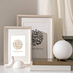 Könnte beinhalten: Zwei gerahmte Drucke mit arabischer Kalligraphie in Gold und Schwarz. Die Drucke sind in weißen Rahmen und haben den Text "O Allah Bless This House" in Englisch unter der Kalligraphie.