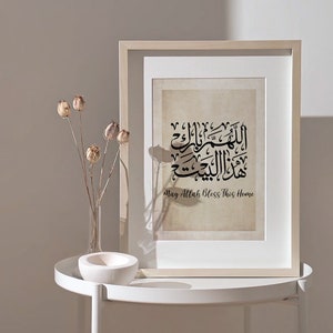 Op de afbeelding: Een ingelijst afdruk met een beige achtergrond en zwarte Arabische kalligrafie. De tekst luidt "Moge Allah dit huis zegenen". De afdruk wordt weergegeven op een witte tafel met gedroogde bloemen in een glazen vaas.