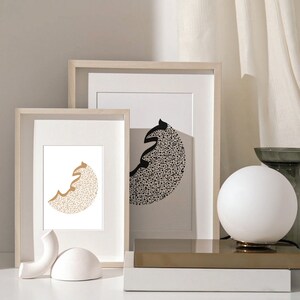 May include: Two framed prints with black and gold abstract designs. The prints are in simple white frames and are displayed on a white surface.