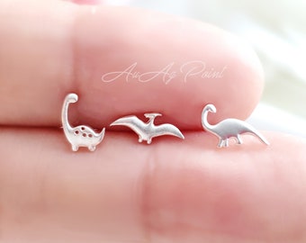 May include: Three silver dinosaur earrings, one is a pterodactyl, one is a long-necked dinosaur, and the other is a traditional dinosaur. The earrings are small and delicate.