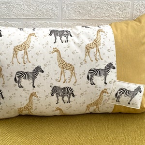 May include: A decorative cushion with a white background and a pattern of giraffes and zebras in black, white, and brown. The cushion has a mustard yellow border and a small mustard yellow pocket with a zebra pattern on the side.