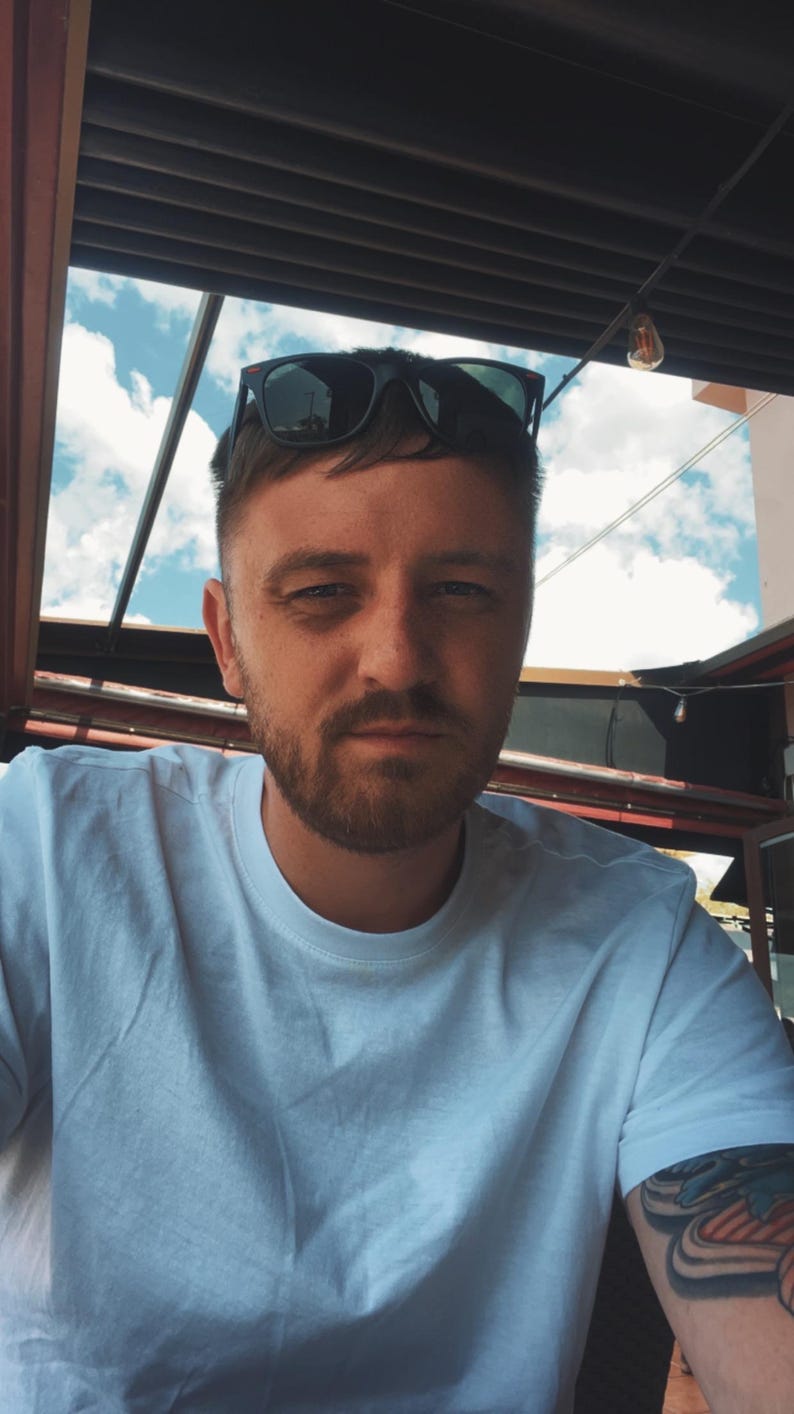 May include: A man wearing a white t-shirt and black sunglasses. The man has short brown hair and a beard. The background shows a blue sky with clouds and a partial view of a building.