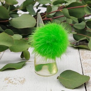 Könnte beinhalten: Ein flauschiger Pompon in leuchtendem Grün auf einem transparenten Präsentationsständer. Der Pompon besteht aus Kunstfell und hat einen Durchmesser von etwa 5 cm.