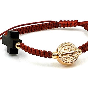 May include: Red braided bracelet with a gold-toned Saint Benedict medal and a black wooden cross charm.