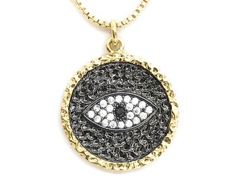 May include: Gold-toned necklace featuring a circular pendant with an eye design. The pendant has a textured black background, a gold-colored textured border, and an eye made of white and black stones. The chain is a delicate gold color.
