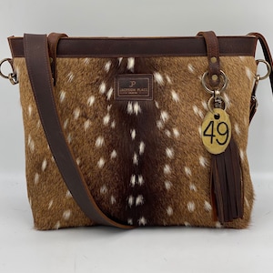 May include: A brown leather handbag with a white and brown spotted pattern. The bag has a long brown leather strap and a brass key chain with a tag that says "49". The bag has a label that says "P Jackson Place".