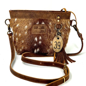 Puede incluir: Bolso bandolera marrón y blanco moteado con correa de cuero. El bolso tiene forma rectangular con una parte superior de cuero marrón y una pequeña etiqueta con el número 39. El bolso tiene una borla y herrajes metálicos.