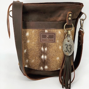 May include: A brown leather shoulder bag with a deer hide front panel. The bag features a long adjustable strap, a metal tag with the number 33, and a leather tassel. The bag has the text "JACKSON PLACE" on a leather patch.