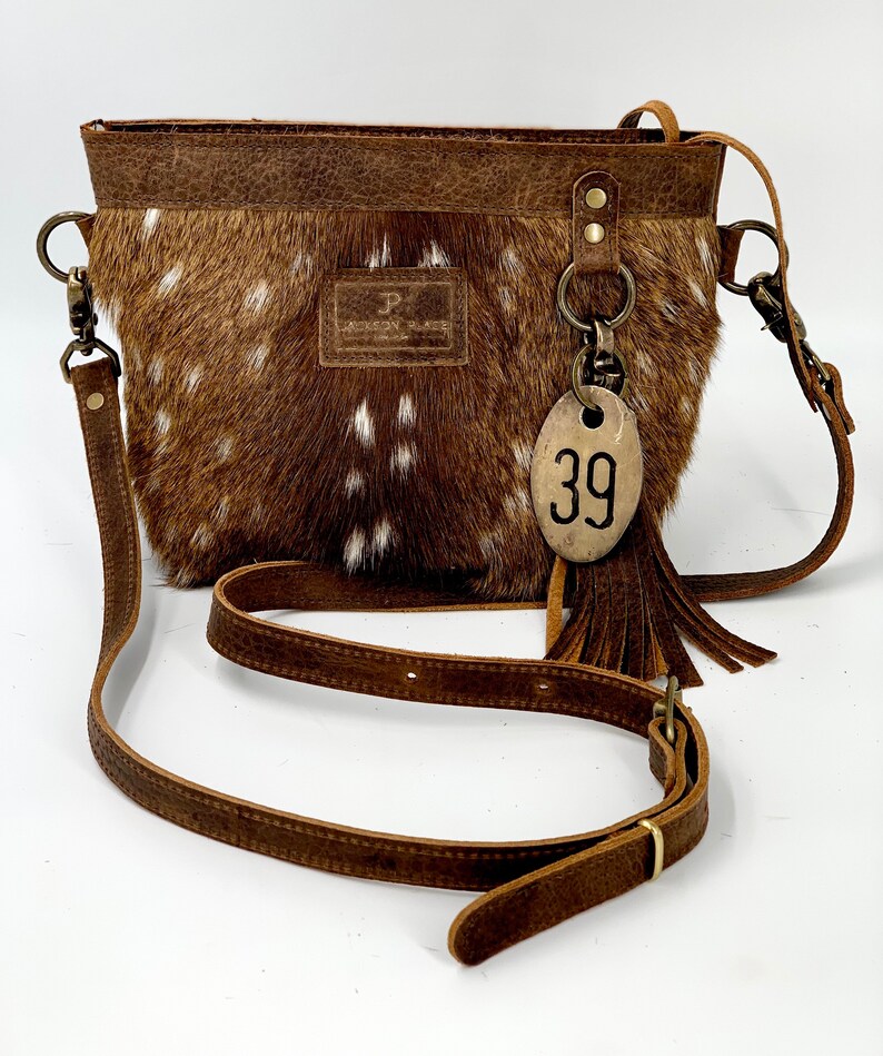 Puede incluir: Bolso bandolera marr&oacute;n y blanco con estampado de lunares y correa de cuero. El bolso tiene forma rectangular con la parte superior de cuero marr&oacute;n y una peque&ntilde;a etiqueta con el n&uacute;mero 39. Tiene una borla y una peque&ntilde;a etiqueta de cuero con la letra "P".