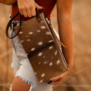 May include: A brown and white spotted leather wallet with a wrist strap. The wallet has a zipper closure and a brown leather strap. The background is blurred, with tall, dry grass.