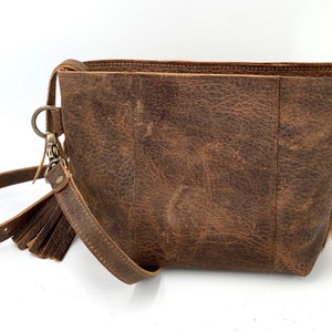 Puede incluir: Bolso bandolera de cuero marr&oacute;n con textura envejecida. El bolso cuenta con una correa de mu&ntilde;eca desmontable con borla y un cierre de metal plateado. El bolso tiene forma rectangular y un dise&ntilde;o sencillo.