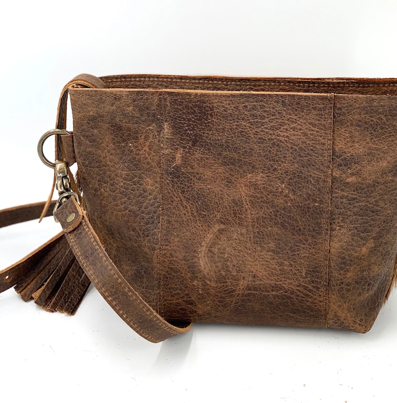 Puede incluir: Bolso bandolera de cuero marr&oacute;n con textura envejecida. El bolso cuenta con una correa de mu&ntilde;eca desmontable con borla y un cierre de metal plateado. El bolso tiene forma rectangular y un dise&ntilde;o sencillo.