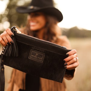 Puede incluir: Clutch de cuero negro con una correa larga y un borla. El clutch tiene una pequeña etiqueta rectangular con el texto "Jackson Place" y un logotipo "J" y "P".