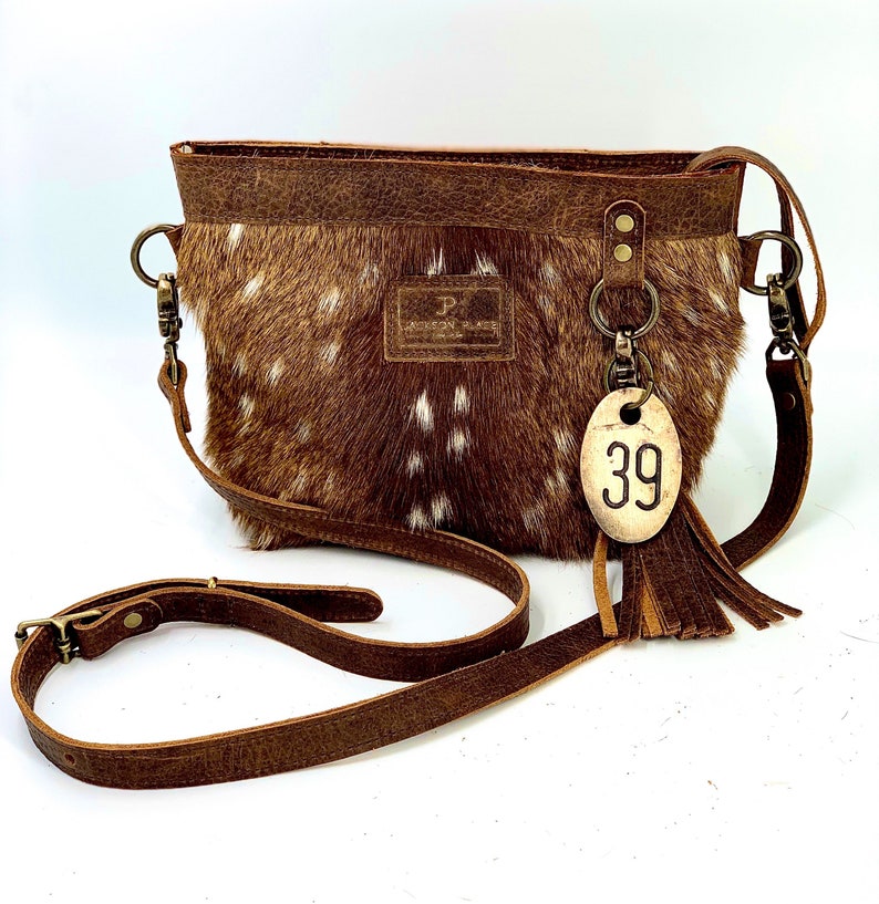 Puede incluir: Bolso bandolera marr&oacute;n y blanco moteado con correa de cuero. El bolso tiene una parte superior de cuero marr&oacute;n y una etiqueta con el n&uacute;mero 39. El bolso tiene una borla y un cierre met&aacute;lico.