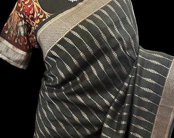 May include: Black saree with silver-colored diagonal stripes and a decorative border. The saree features a patterned blouse with red, yellow, and brown accents. A diamond necklace is also visible.
