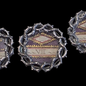Pode incluir: Três broches circulares em estilo antigo com uma borda prateada de videira espinhosa. O centro de cada broche apresenta um fundo roxo com detalhes dourados e o numeral romano VIII.