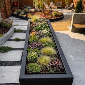 Puede incluir: Diseño de jardín moderno con una jardinera negra rectangular y larga llena de varias plantas y pastos. Un camino de losas cuadradas conduce al jardín, con una terraza de madera y una zona de estar en el fondo.