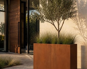 May include: A rectangular, rust-coloured metal planter box containing a small olive tree and tufts of green ornamental grass. The planter has a smooth, weathered finish and sits on a paved surface, showcasing a clean, geometric design.