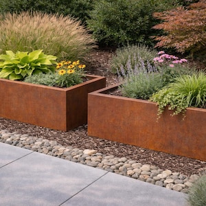 Puede incluir: Dos jardineras rectangulares de metal color óxido llenas de varias plantas verdes y flores. Las jardineras se asientan sobre un lecho de mantillo y pequeñas piedras, con un patio de hormigón gris en primer plano. El fondo presenta una exuberante vegetación.