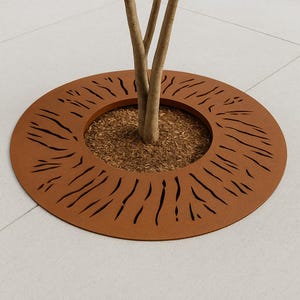 May include: A circular, rust-colored metal tree ring with a series of cut-out lines. The ring surrounds the base of a tree, which is surrounded by mulch. The tree ring sits on a light-colored, tiled surface.