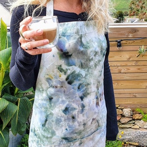 Peut inclure: Une personne portant un tablier tie-dye bleu et vert avec une poche. Elle tient un verre de café avec une couche de mousse sur le dessus.