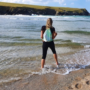 May include: A woman wearing black pants and a black shirt stands in the shallow water of a beach. She is wearing a green and white drawstring bag. The beach is sandy with footprints and the ocean is blue and clear.