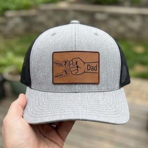 May include: A gray and black trucker hat with a leather patch. The patch features two fists bumping, with the word "Dad" and the names "Prentiss" and "Harvey". The hat has a curved brim and a mesh back.