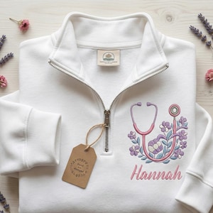 May include: White quarter-zip sweatshirt with a pink stethoscope and floral embroidery. The name "Hannah" is embroidered in pink script below the stethoscope design. A brown tag with the words "Hand stamped" hangs from the zipper.