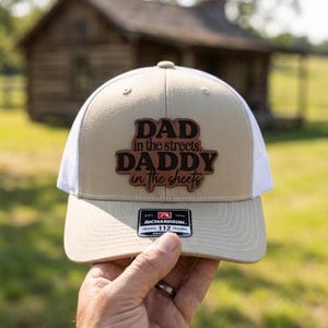 May include: Tan and white trucker hat with the text "DAD in the streets, DADDY in the sheets" in brown lettering. The hat has a curved brim and a mesh back. The Richardson brand tag is visible.