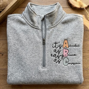 May include: A gray quarter-zip sweatshirt with the text "it's as easy as ABC" in black script. The letters A, B, and C are in orange, pink, and green, respectively, with the words "Antecedent", "Behavior", and "Consequence" next to them.