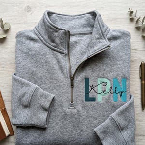 May include: A gray quarter-zip sweatshirt with the embroidered text "LPN Kelly" in teal and light blue. The sweatshirt is folded neatly on a light wood surface, with a pen and notebook visible.