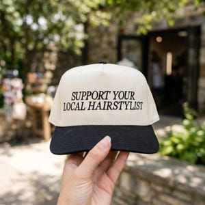 May include: A cream-colored baseball cap with a black brim. The cap features the text "SUPPORT YOUR LOCAL HAIRSTYLIST" in black, centered on the front. The hat is held up in front of a blurred outdoor background.