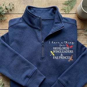 May include: Navy blue quarter-zip sweatshirt with embroidered text and graphics. The text reads "I have a thing for HIGHLORDS WINGLEADERS & FAE PRINCES" with colorful wing graphics. The sweatshirt has a high collar and a silver zipper.