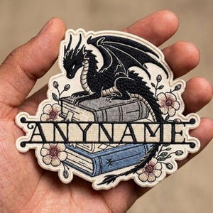 May include: Embroidered patch featuring a black dragon atop two books, with the word "ANYNAME" across the bottom. The patch has a cream-colored border with floral accents in pink and green. The dragon has detailed wings and scales.